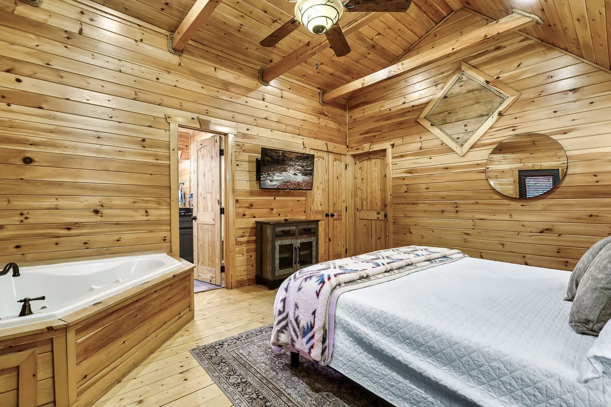 A warmly lit cabin bedroom featuring a wooden bathtub next to a bed, accented by a cozy fireplace and rustic wood-paneled walls. Decorative elements like a mirror and soft linens create an inviting, tranquil atmosphere.