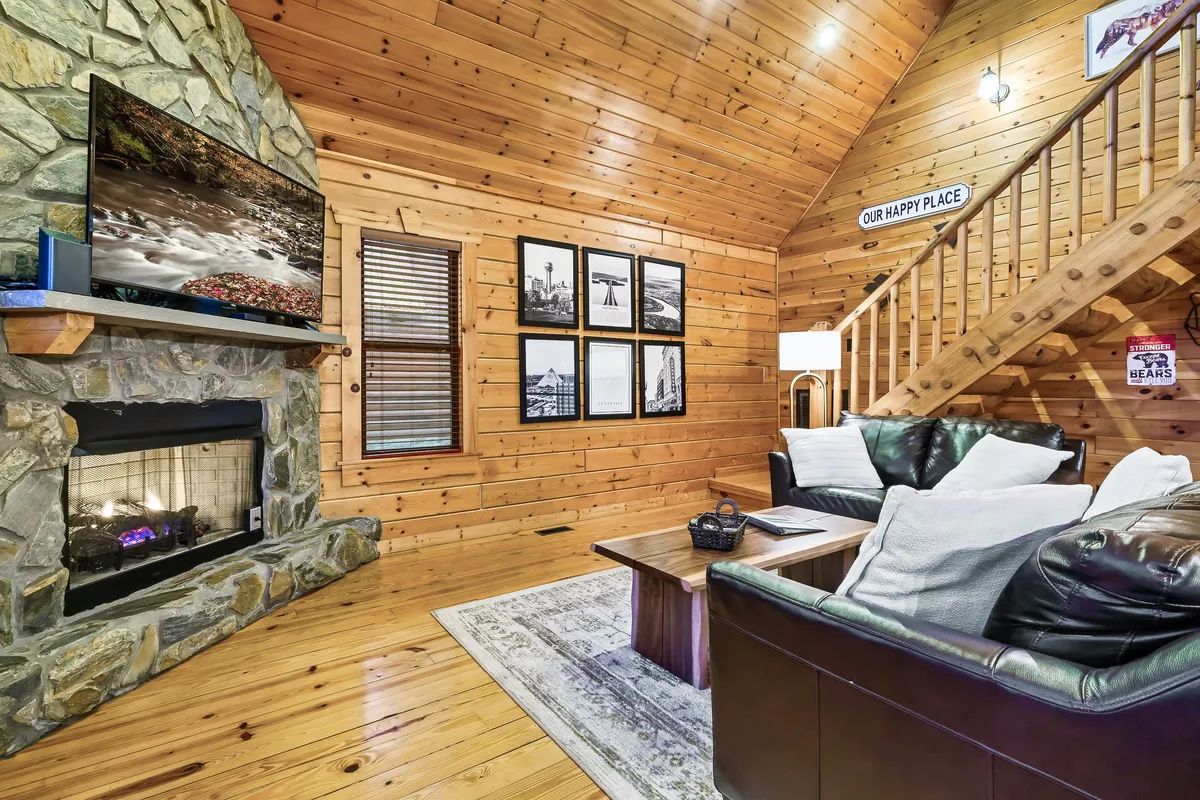 This inviting living area features a substantial stone fireplace as the centerpiece, complemented by a large TV, wooden staircase, photographic wall art, and comfortable seating. The space is open and ideal for group gatherings, cozy evenings, or movie nights.