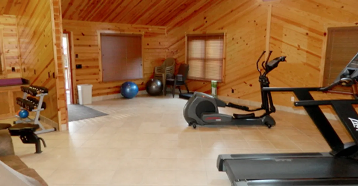 A modern, airy fitness room with exercise equipment, warm wooden paneling, and generous natural light streaming through large windows. The space presents a motivational, serene environment ideal for personal fitness and wellness routines.