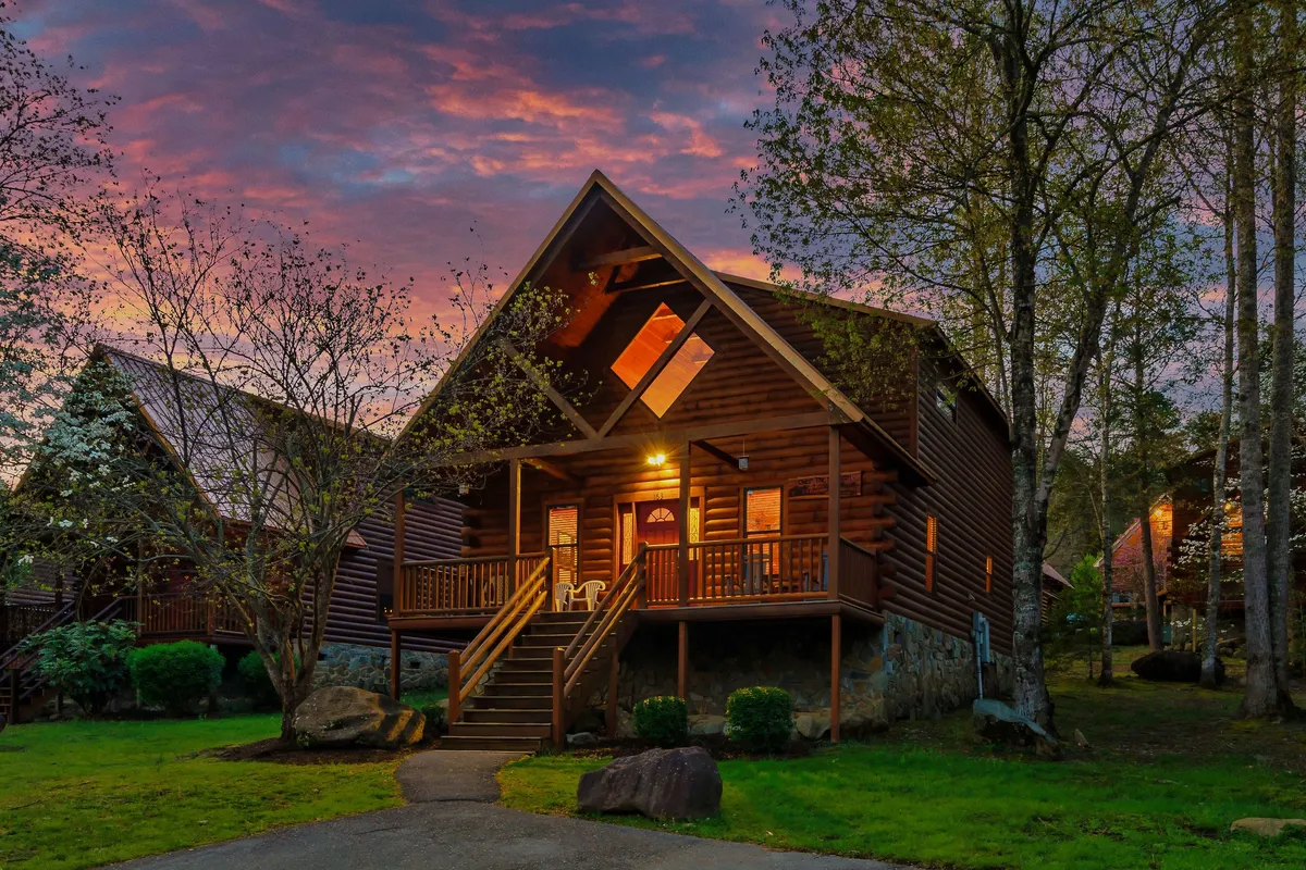 The log cabin's exterior with wide steps leading up to a welcoming front porch, set among tall trees. Large windows hint at the spacious interior, while the inviting porch offers a glimpse of an ideal spot for outdoor meals or relaxed group conversations.