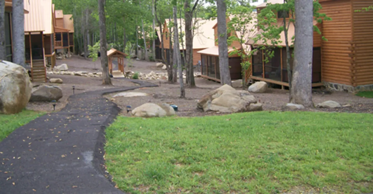 A serene pathway winding between wooden cabins and large boulders, flanked by trees and open space, ideal for safe family walks, nature scavenger hunts, or easy access to resort amenities.