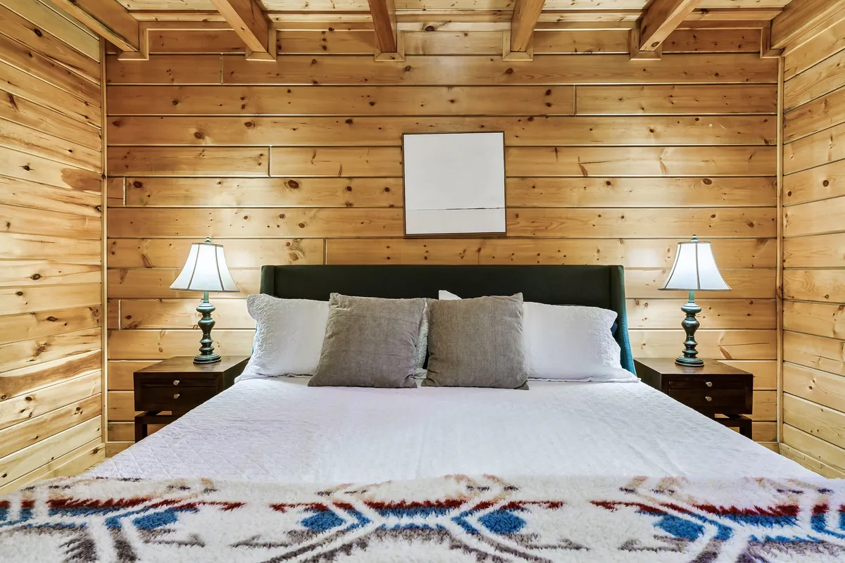 A cozy bedroom with a comfortable bed, wooden walls, matching nightstands, and soft lighting from lamps. The inviting blanket and warm wooden finishes create an atmosphere ideal for relaxation and restful sleep.