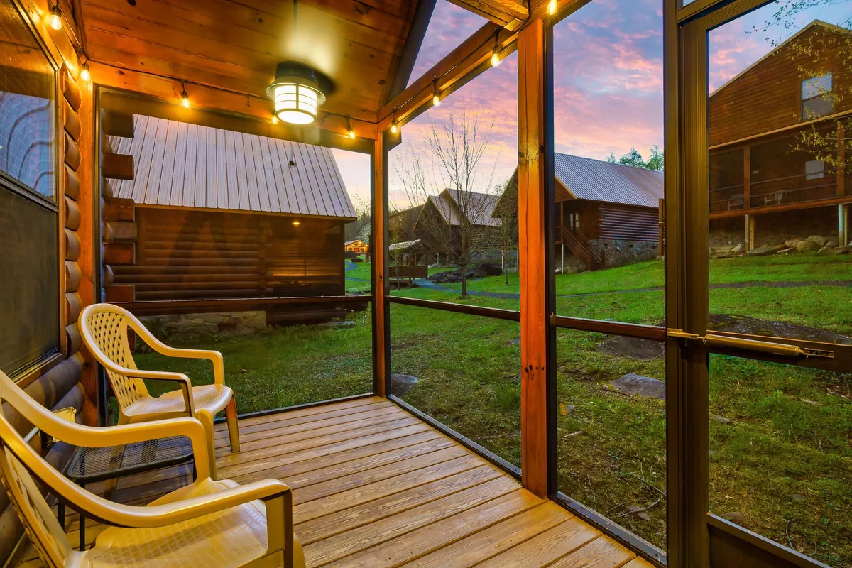 A cozy screened porch framed by wooden beams looks out onto a green lawn. A hanging light fixture provides inviting evening ambiance, while the serene setting suggests moments of relaxation after an active day outdoors.