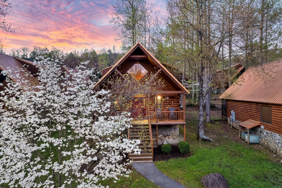 A log cabin framed by flowering trees, a spacious front porch, and vibrant outdoor scenery. The setting is peaceful yet inviting, ideal for relaxing, playing outdoors, or enjoying nature with the family.