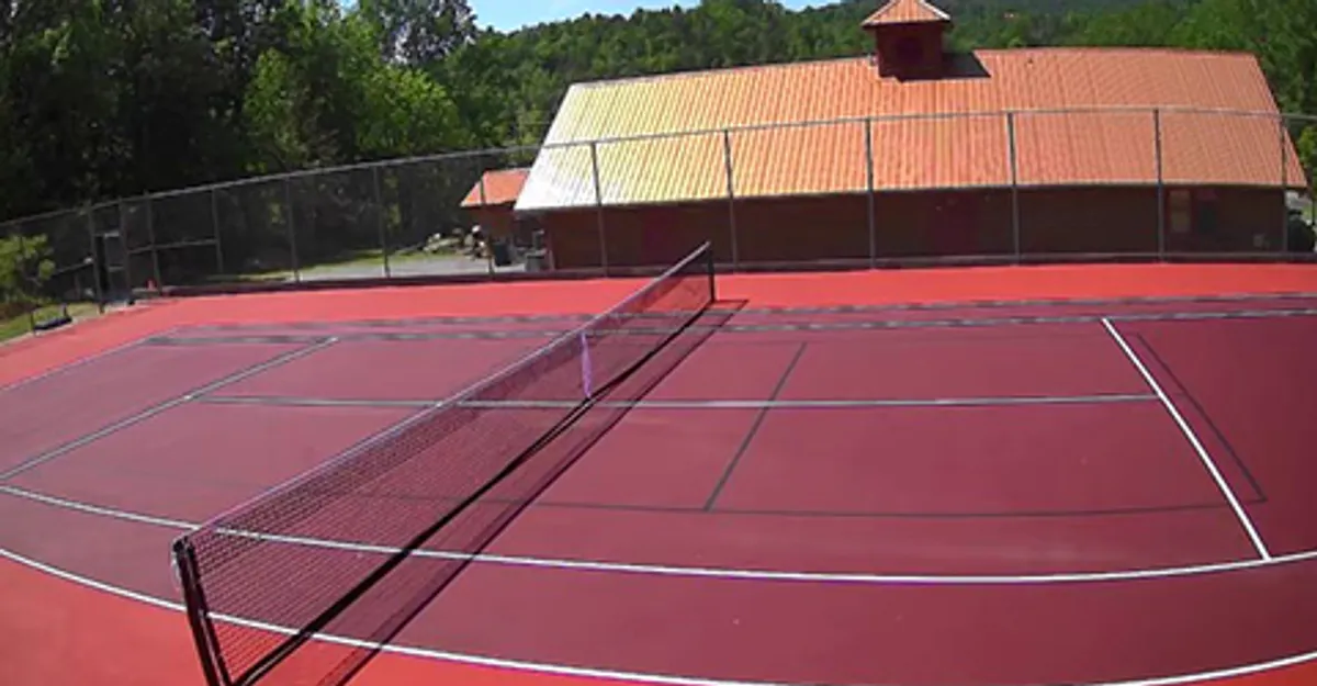 A vibrant red tennis court with a net in the foreground and a resort building visible behind. The scene suggests opportunities for active, social recreation, such as playing pickleball or tennis with friends or family.