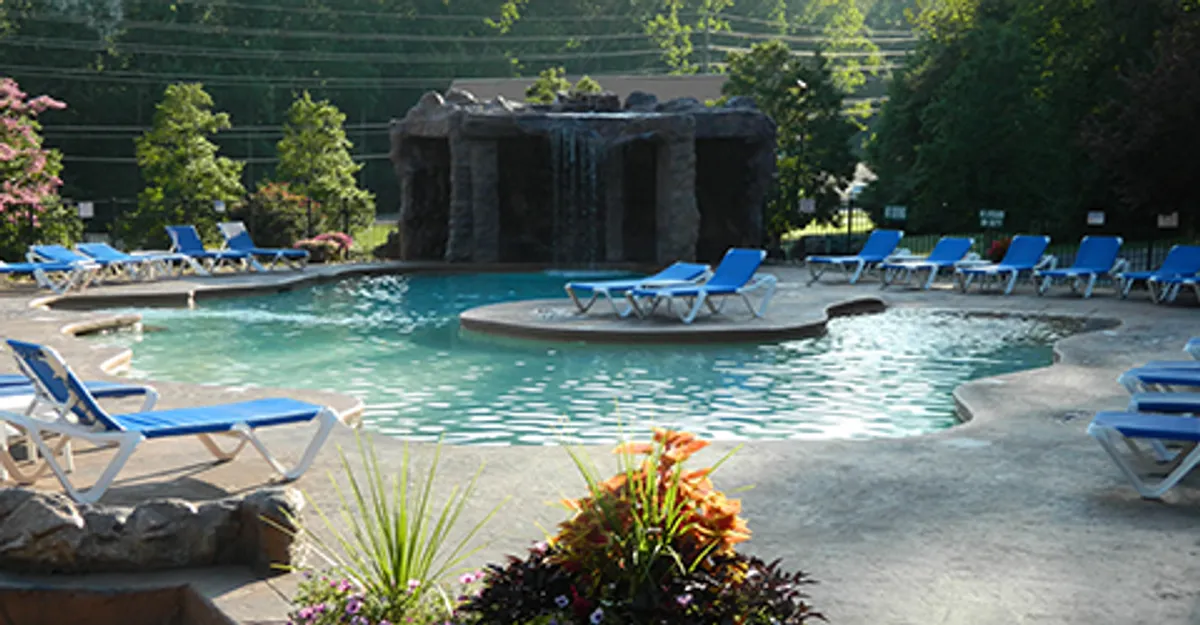 An exterior scene of a sparkling resort-style swimming pool with a picturesque waterfall, surrounded by lush greenery, colorful flowers, and comfortable lounge chairs. The atmosphere is lively yet peaceful, inviting guests to swim, sunbathe, or socialize by the poolside.