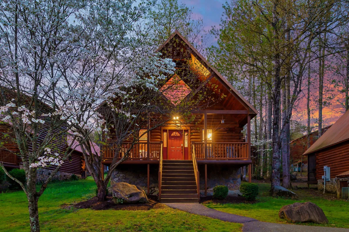 The image showcases a charming wooden cabin nestled among flowering trees, with a welcoming front porch and steps leading up to the door. The cabin is surrounded by lush greenery, hinting at peaceful nature walks and a tranquil atmosphere. The porch invites guests to relax, enjoy sunsets, and gather with loved ones in a cozy, scenic setting.