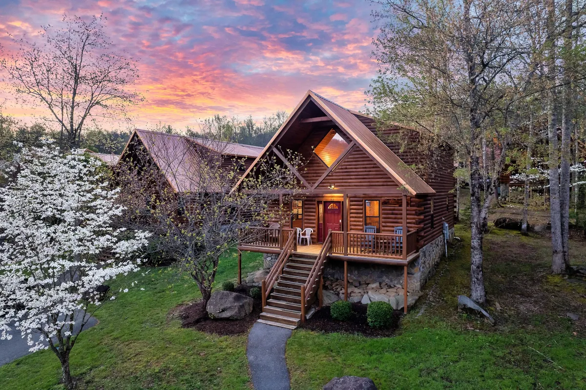 This image captures the exterior of a log cabin set against flowering trees and a vibrant, colorful sunset. The welcoming front porch invites guests to relax, while the backdrop of nature hints at the surrounding mountain scenery and outdoor adventure awaiting just beyond the doorstep.