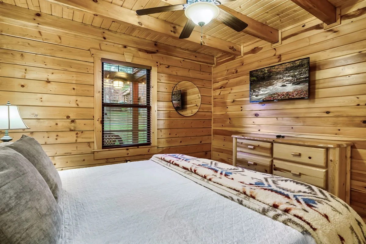 A cozy bedroom with a patterned blanket on the bed, rustic wooden furniture, a round mirror, and framed artwork on the walls. Natural light filters into the inviting, peaceful space.